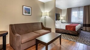 A hotel room with a brown sofa, a small table, a bed with a red blanket, a nightstand with a lamp, and a picture on the wall. - Comfort Inn & Suites Fruita - Grand Junction (Fruita)