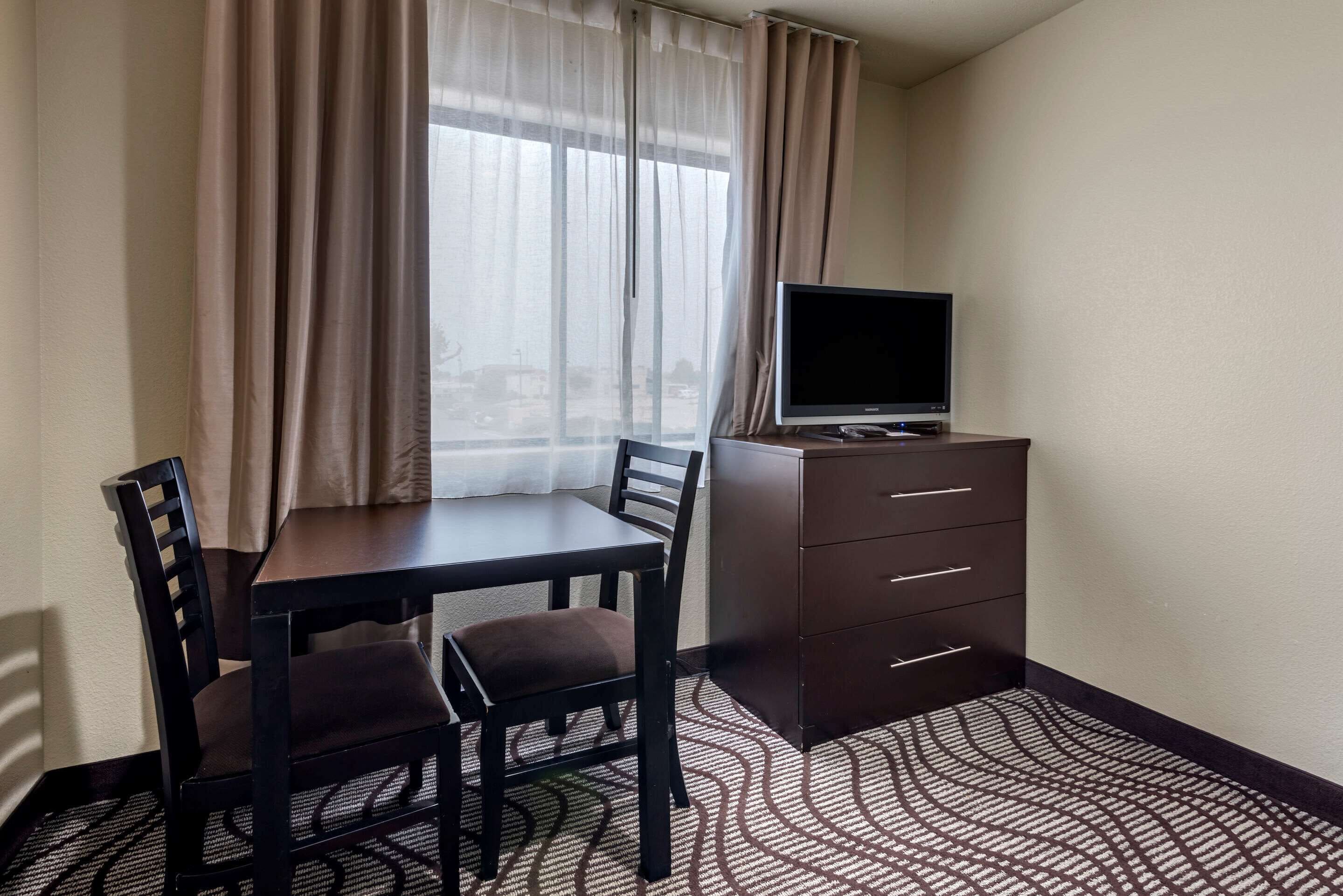 A hotel room with a dining table, two chairs, a TV on a dresser, and a window with curtains.