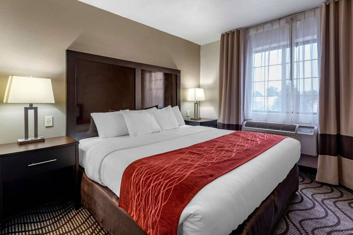 A hotel room with a large bed, two bedside tables with lamps, a window with curtains, and an air conditioning unit.