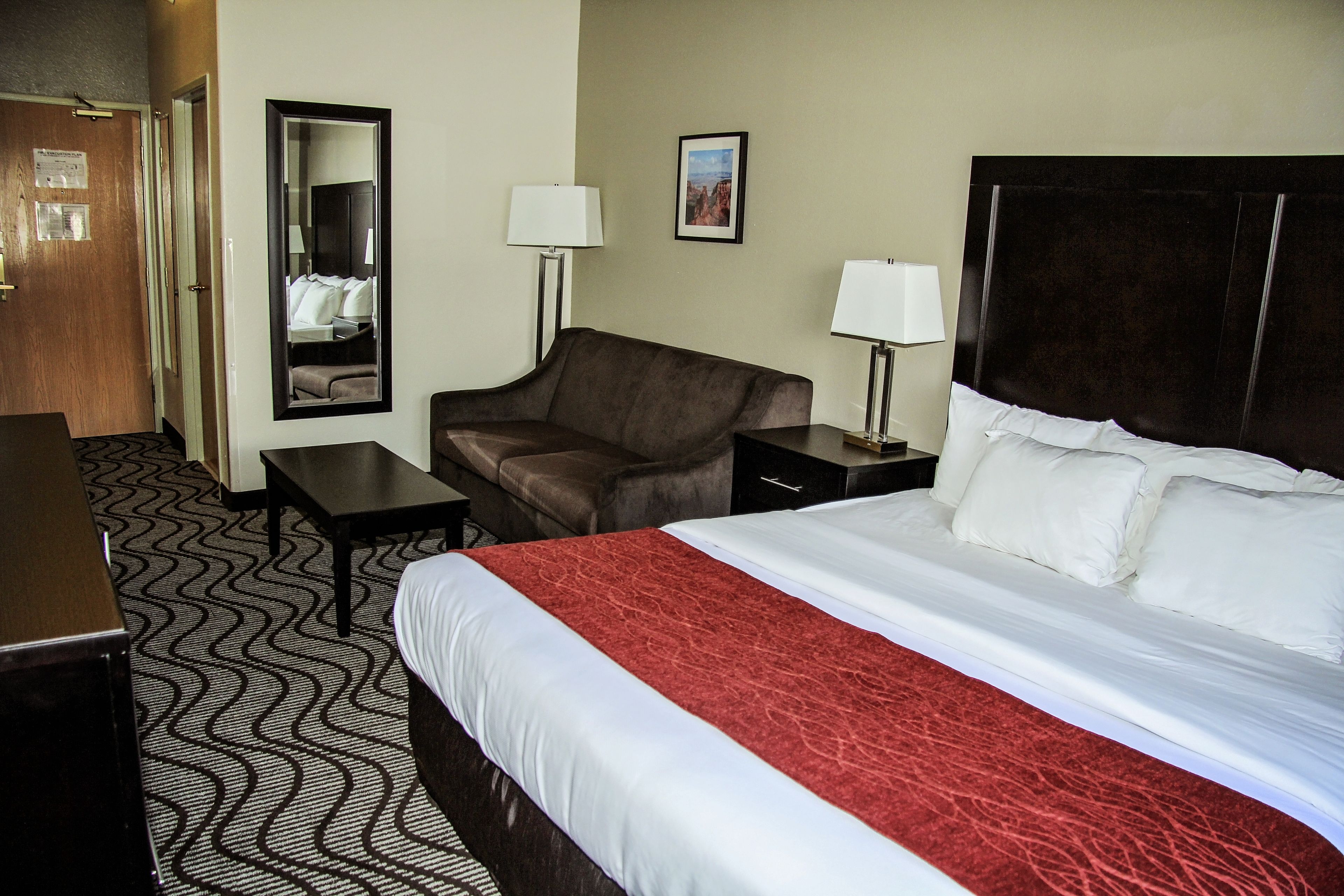 A hotel room with a large bed, a desk, a chair, a mirror, and a nightstand with lamps.