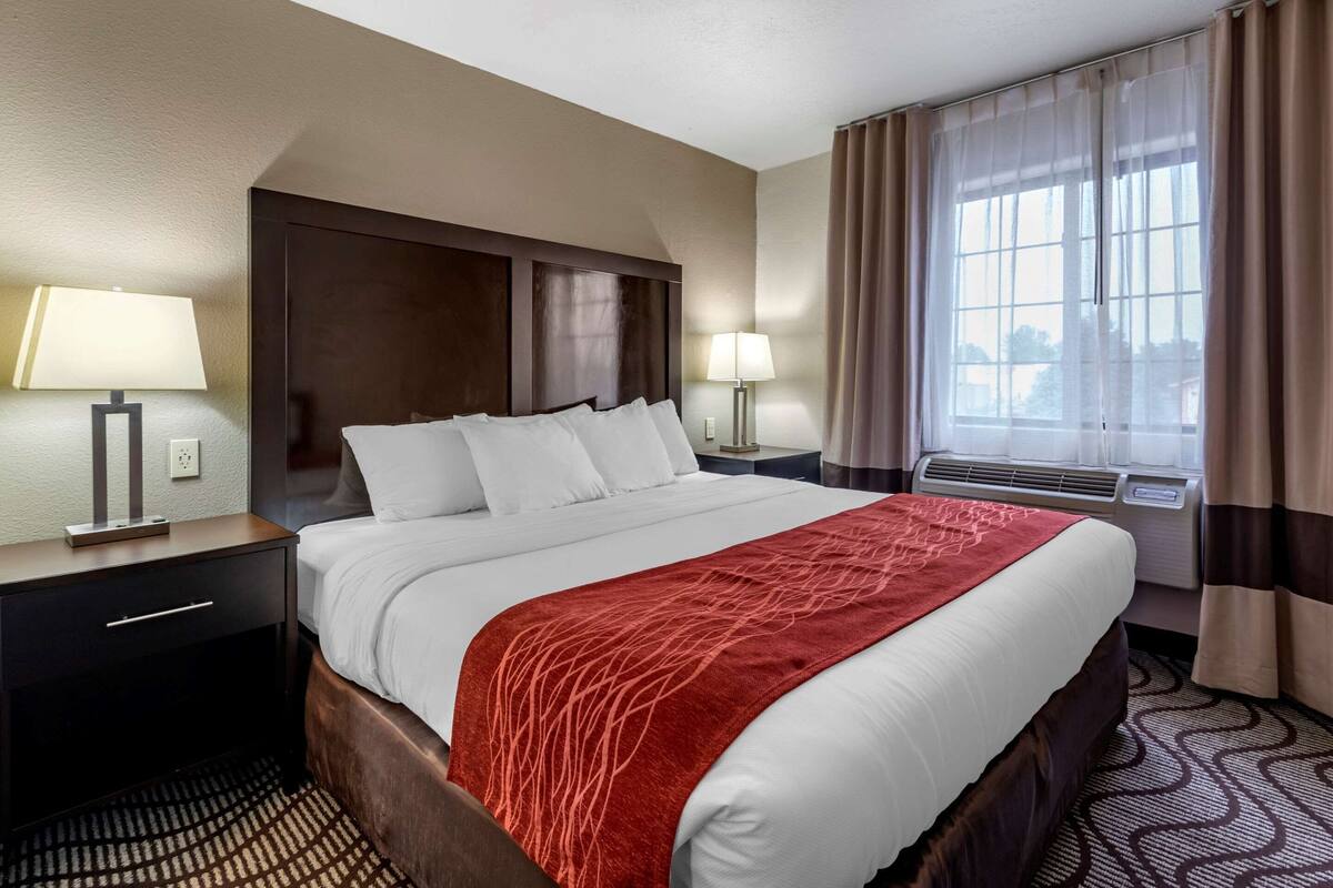 A hotel room with a large bed, two bedside tables with lamps, a window with curtains, and an air conditioning unit.