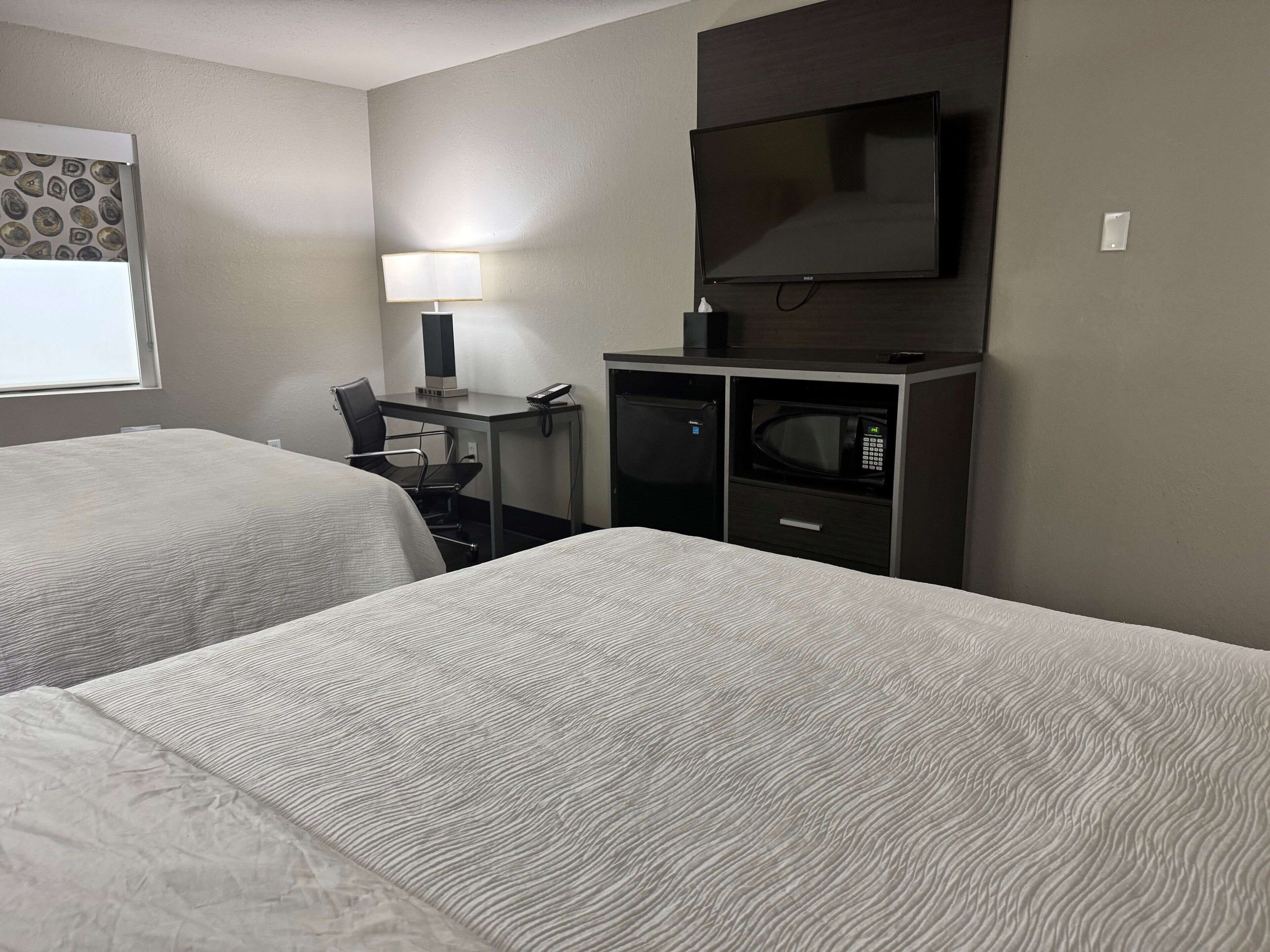 A hotel room with two beds, a desk with a lamp, a TV, a microwave, and a refrigerator.