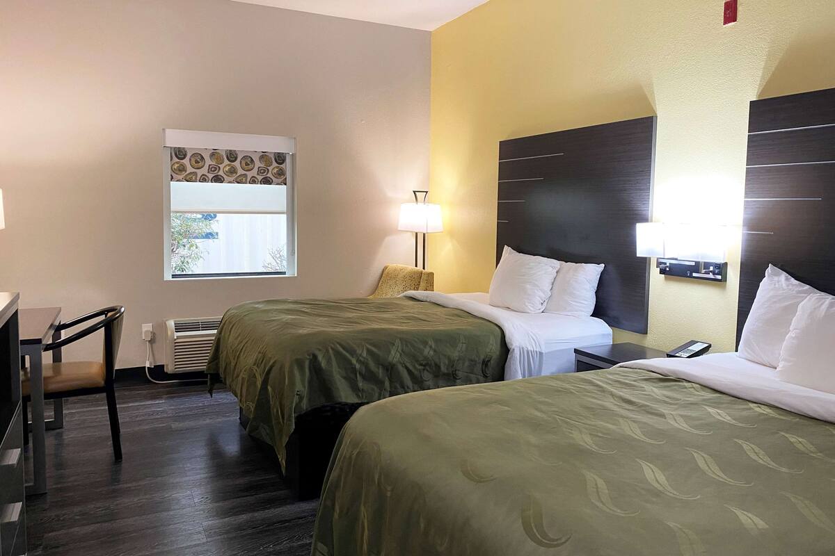 Une chambre d’hôtel avec deux lits, un bureau, une chaise, une fenêtre avec des stores, une lampe et un téléviseur fixé au mur.