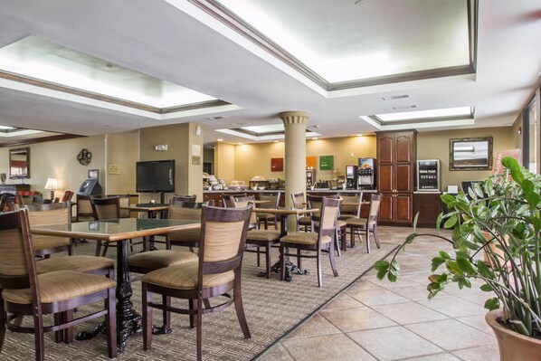 Un vestíbulo de hotel con varias mesas y sillas para comer, un pilar grande, un televisor y una máquina de café.