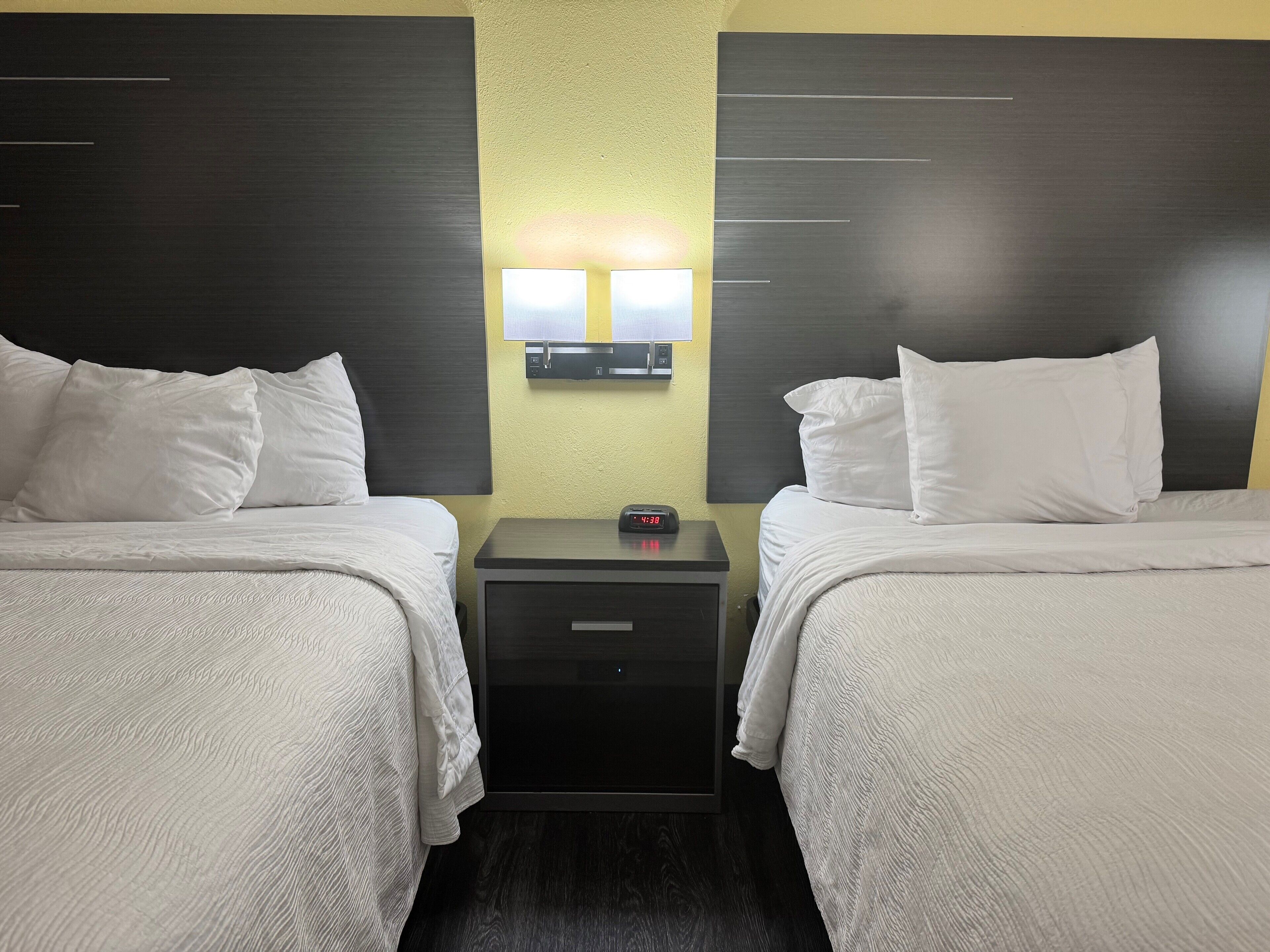 A hotel room with two beds, a nightstand with a clock, and a wall-mounted light fixture.