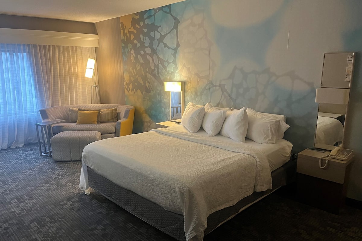 A hotel room with a large bed, a chair, a desk, and a nightstand with a phone.