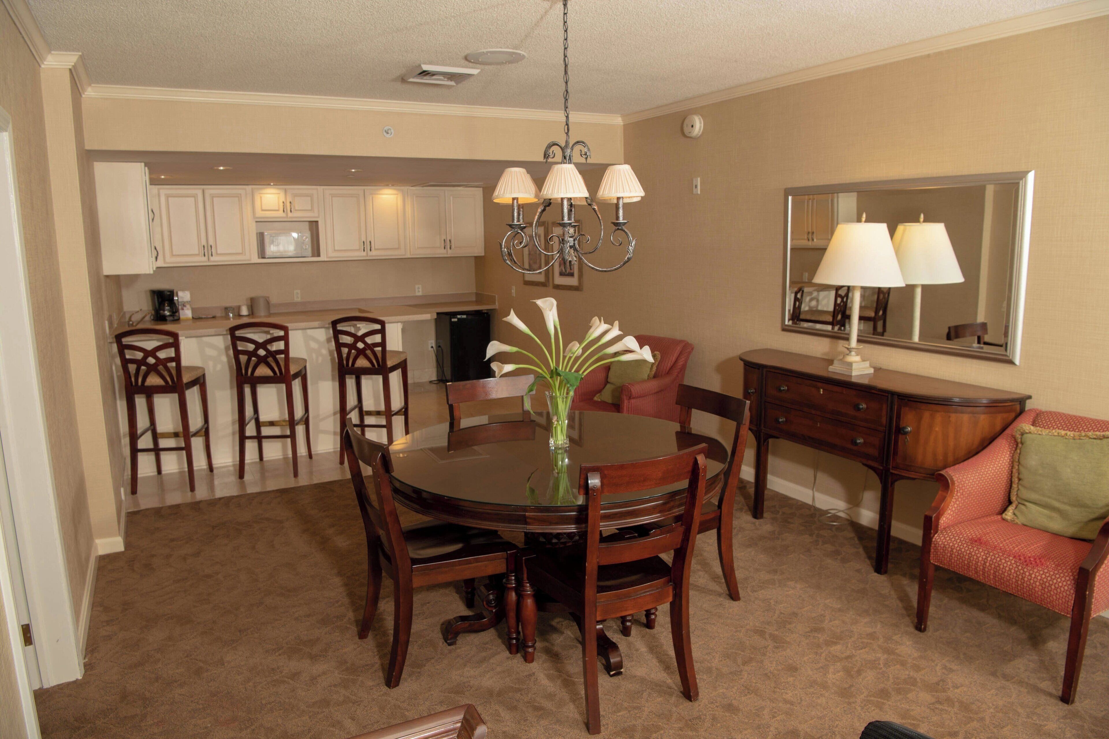 A dining area with a round table, chairs, a vase with flowers, a chandelier, a sofa, a dresser, and a mirror.
