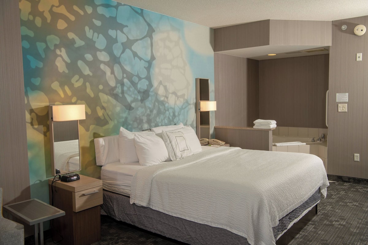 A hotel room with a large bed, bedside tables, a mirror, a wall-mounted phone, and a bathroom visible through an open door.