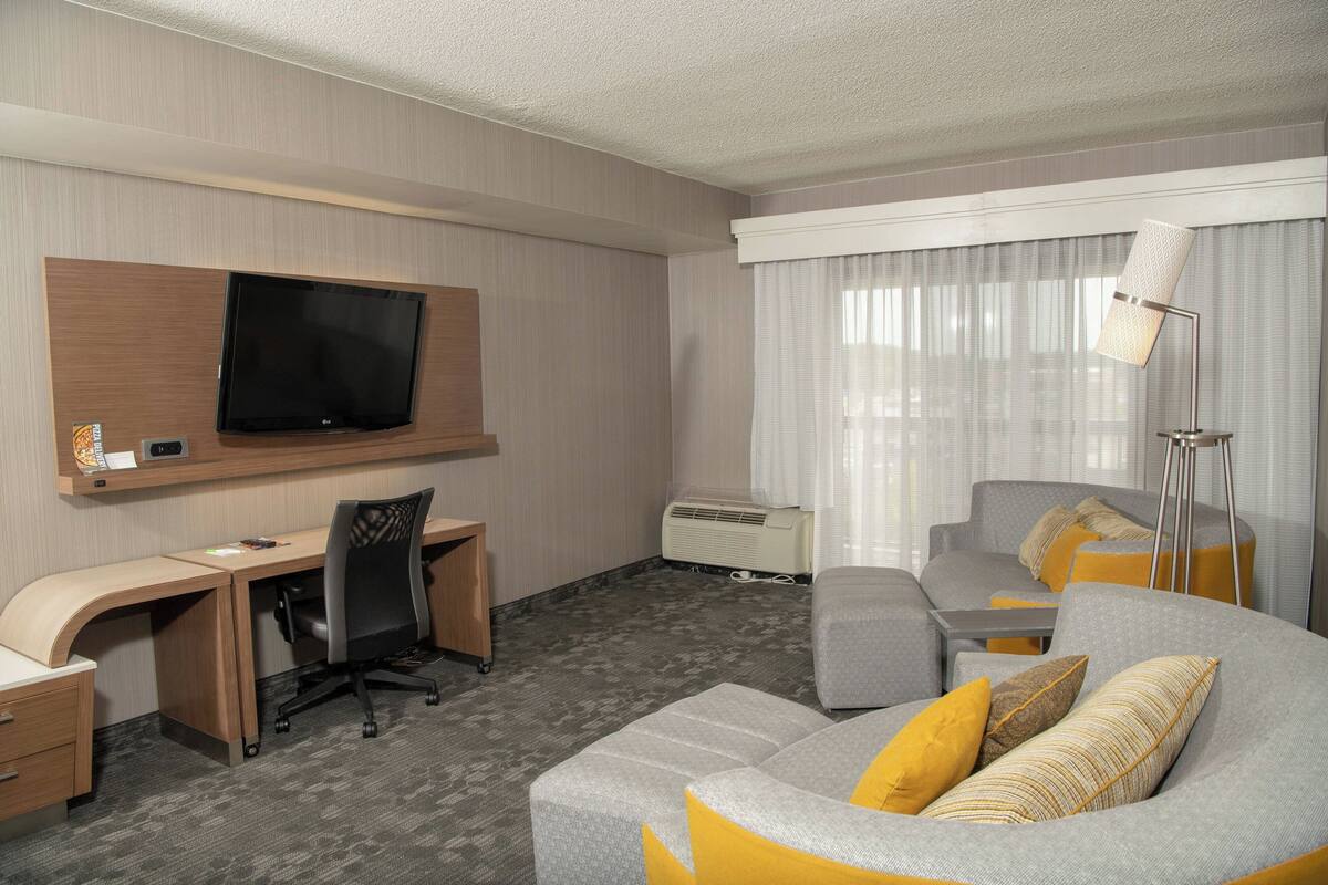 A hotel room with a flat-screen TV mounted on the wall, a desk with a chair, a grey sofa with yellow cushions, and a floor lamp.