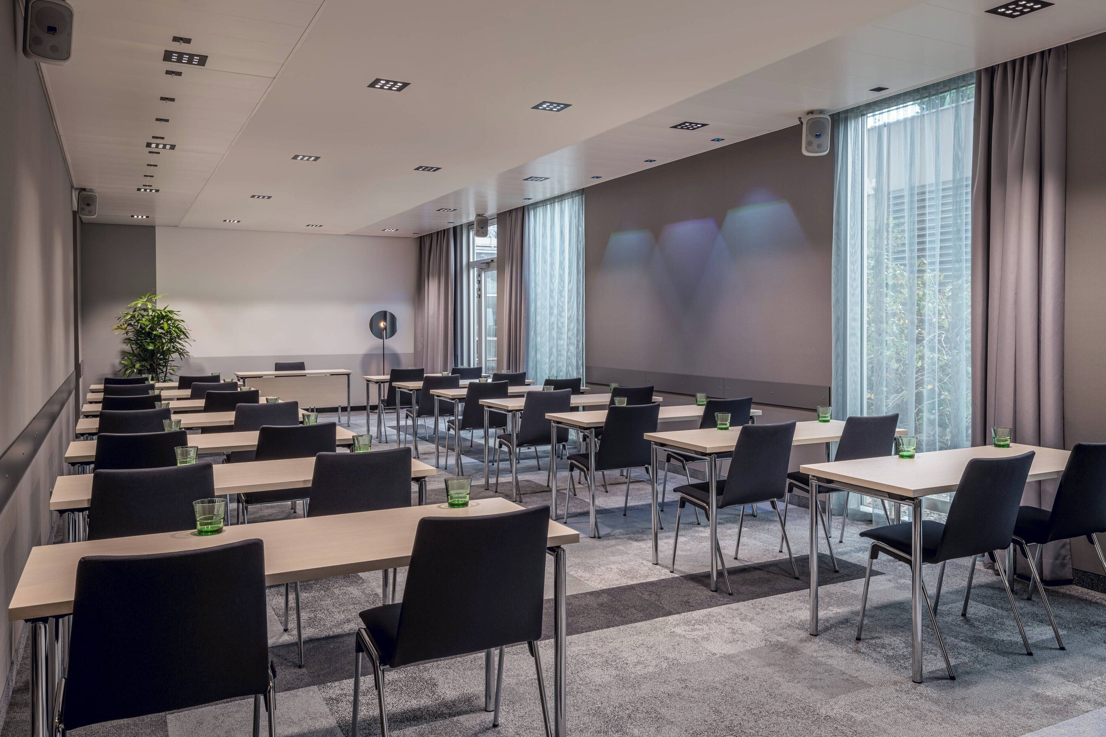 Une salle de conférence équipée de plusieurs tables et chaises, d’un écran de projection et de grandes fenêtres avec des rideaux.