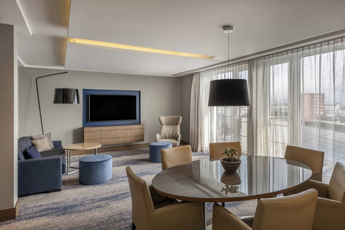 Una habitación de hotel moderna con un televisor de pantalla plana grande, un área de comedor con una mesa redonda y sillas, un sofá, una silla y un amplio ventanal con cortinas.
