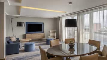 A modern hotel room with a large flat-screen TV, a dining area with a round table and chairs, a sofa, a chair, and a large window with curtains.