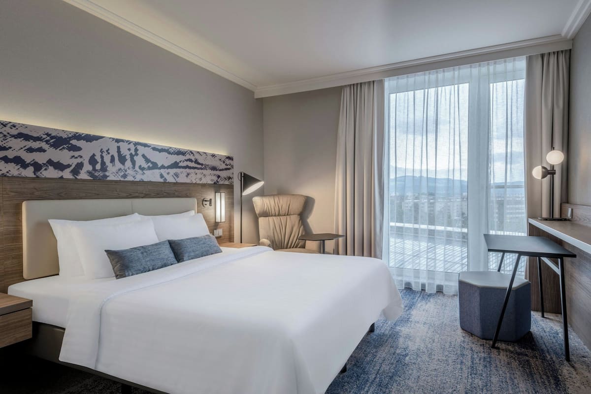 Habitación de hotel con una cama grande, una silla, un escritorio y vistas a la ciudad a través de cortinas traslúcidas.