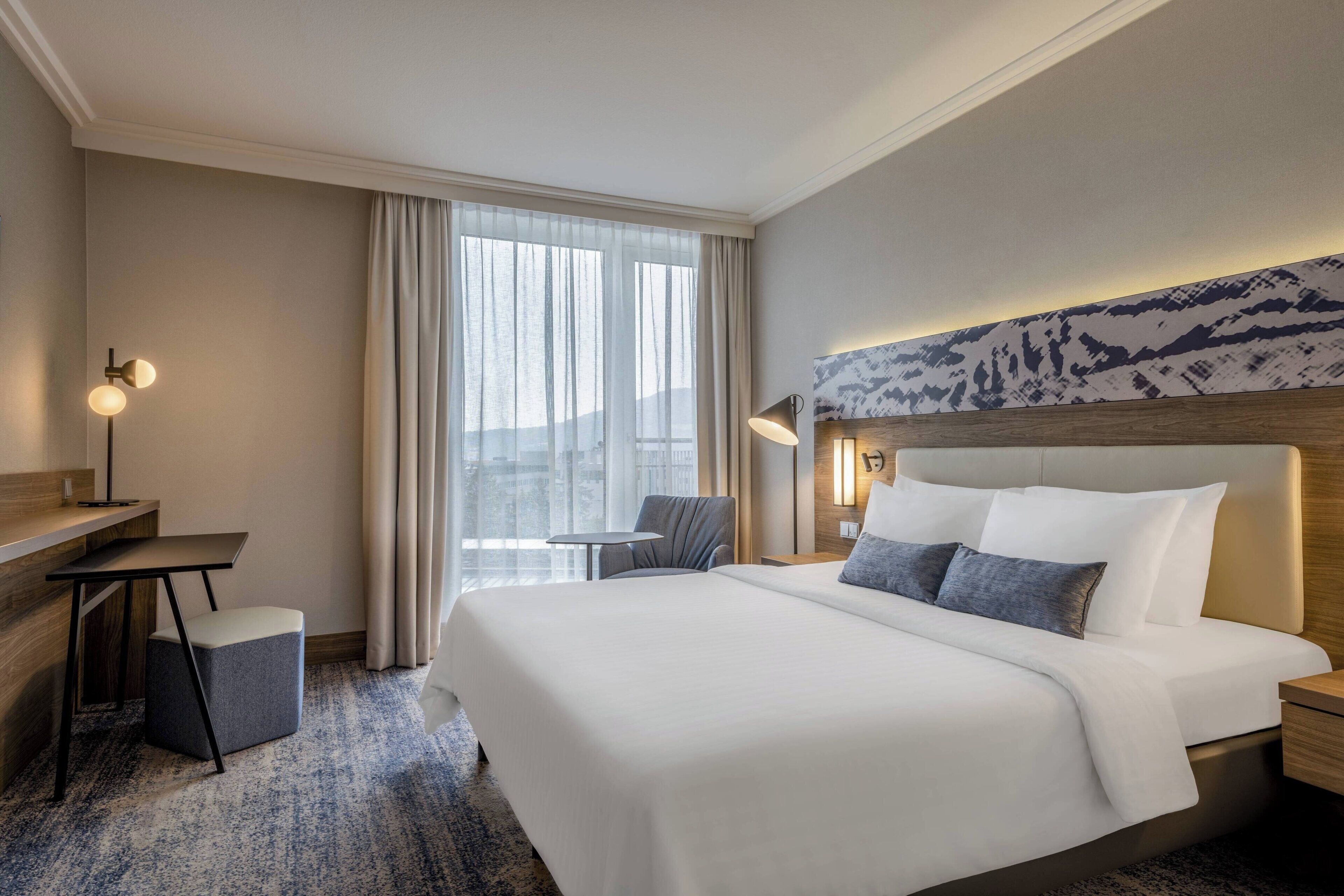 A hotel room with a large bed, a desk, a chair, and a view of the city through the window.