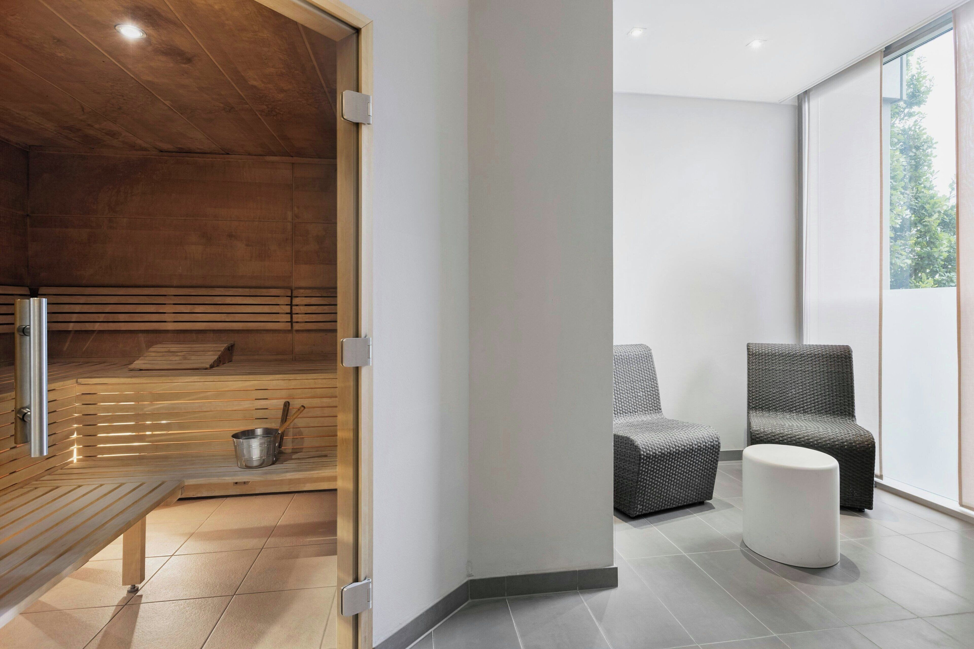 Un área de spa moderna con sauna de madera, un banco, una mesita con una olla y dos sillones grises tapizados.