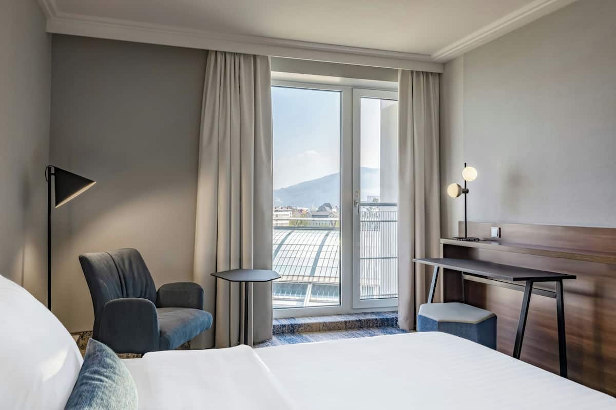Habitación de hotel con una cama grande, un escritorio, una silla y vistas a un paisaje urbano a través de un ventanal amplio.