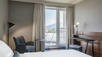 A hotel room with a large bed, a desk, a chair, and a view of a cityscape through a large window.