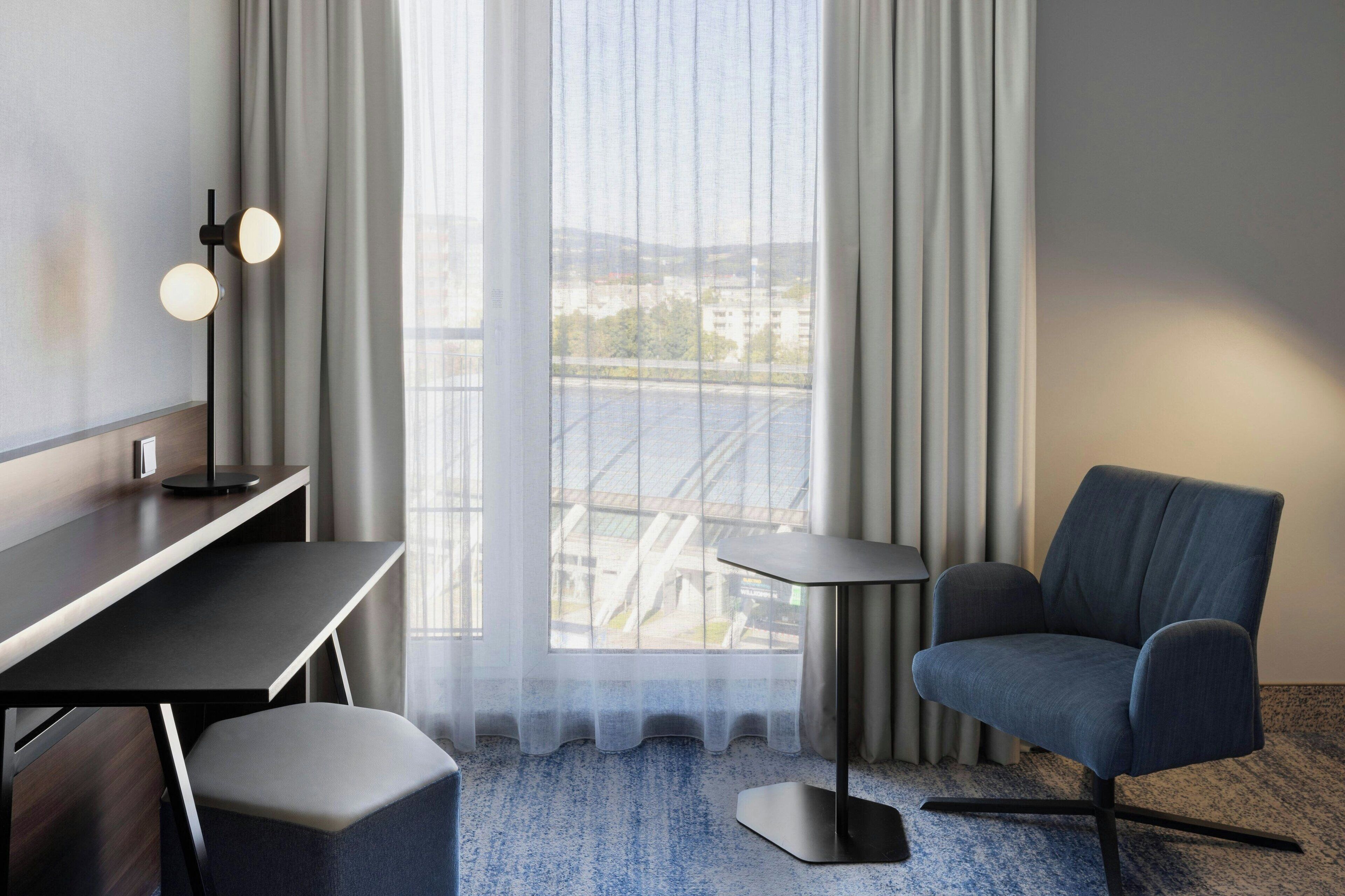 Une chambre d’hôtel moderne, dotée d’un bureau, d’une chaise et offrant une vue sur le paysage urbain grâce à de grandes fenêtres.
