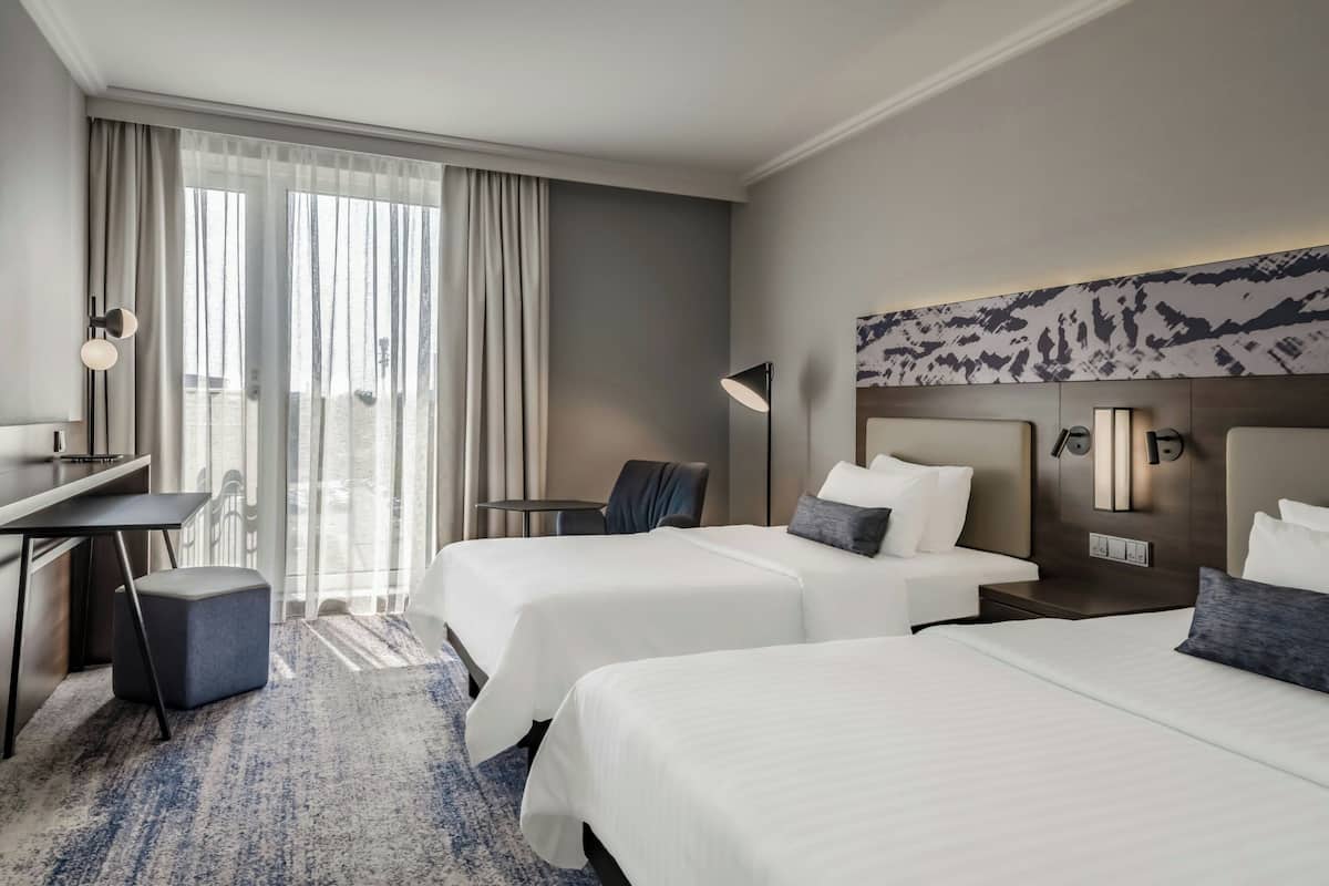 Habitación de hotel con dos camas, un escritorio, una silla y un amplio ventanal con cortinas.