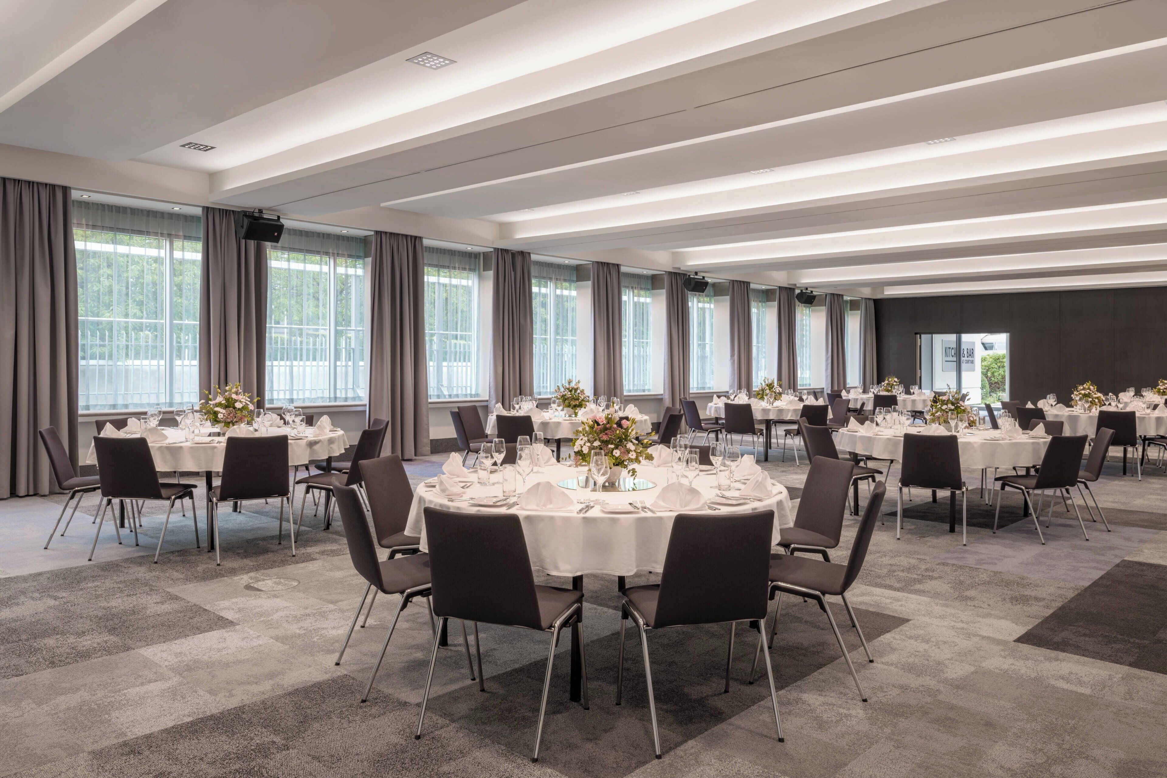 A spacious conference room with multiple round tables set for dining, large windows, and a modern ceiling design.