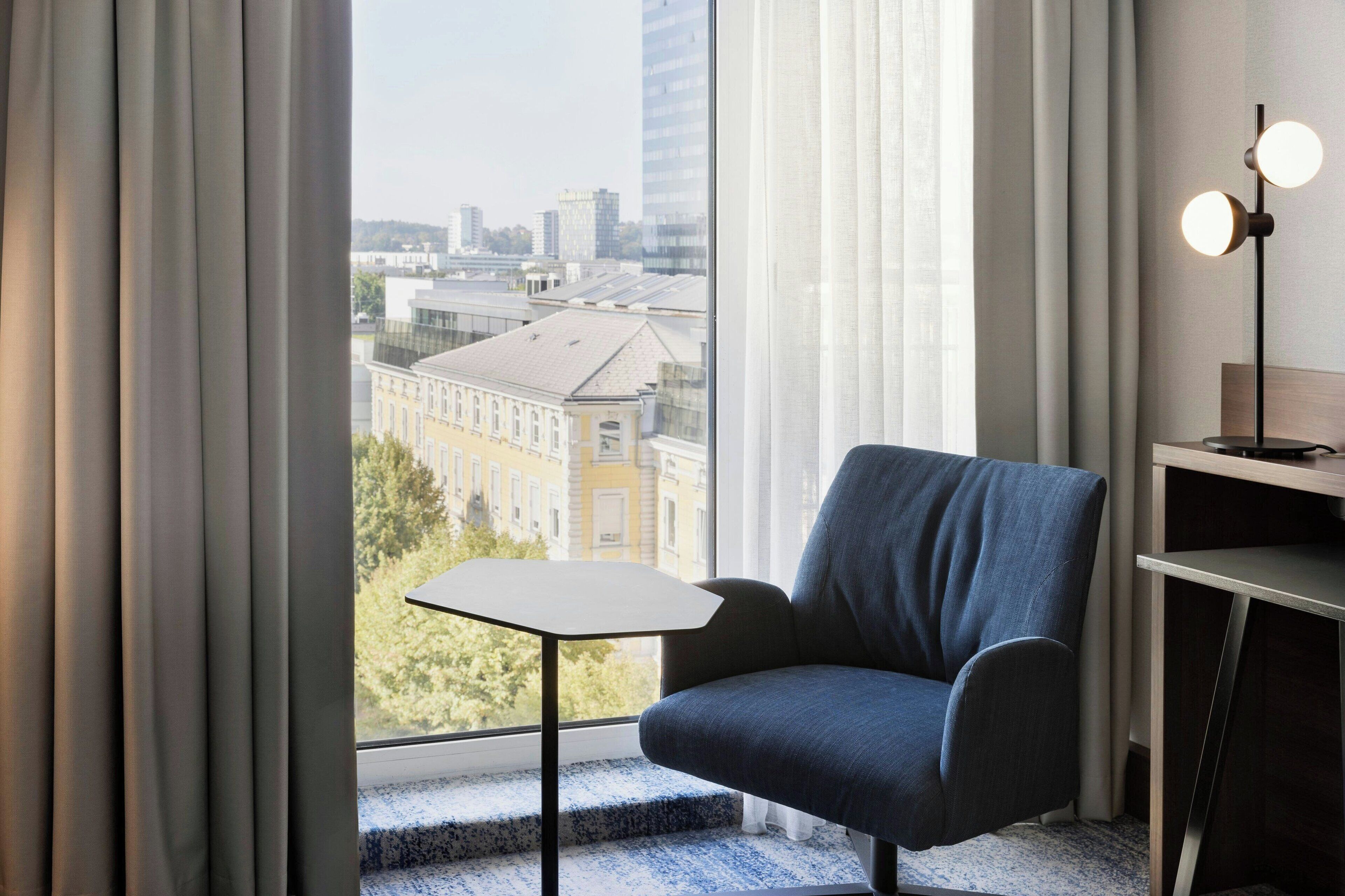 A modern room with a view of a cityscape through large windows, a blue armchair, a small table, and a floor lamp.