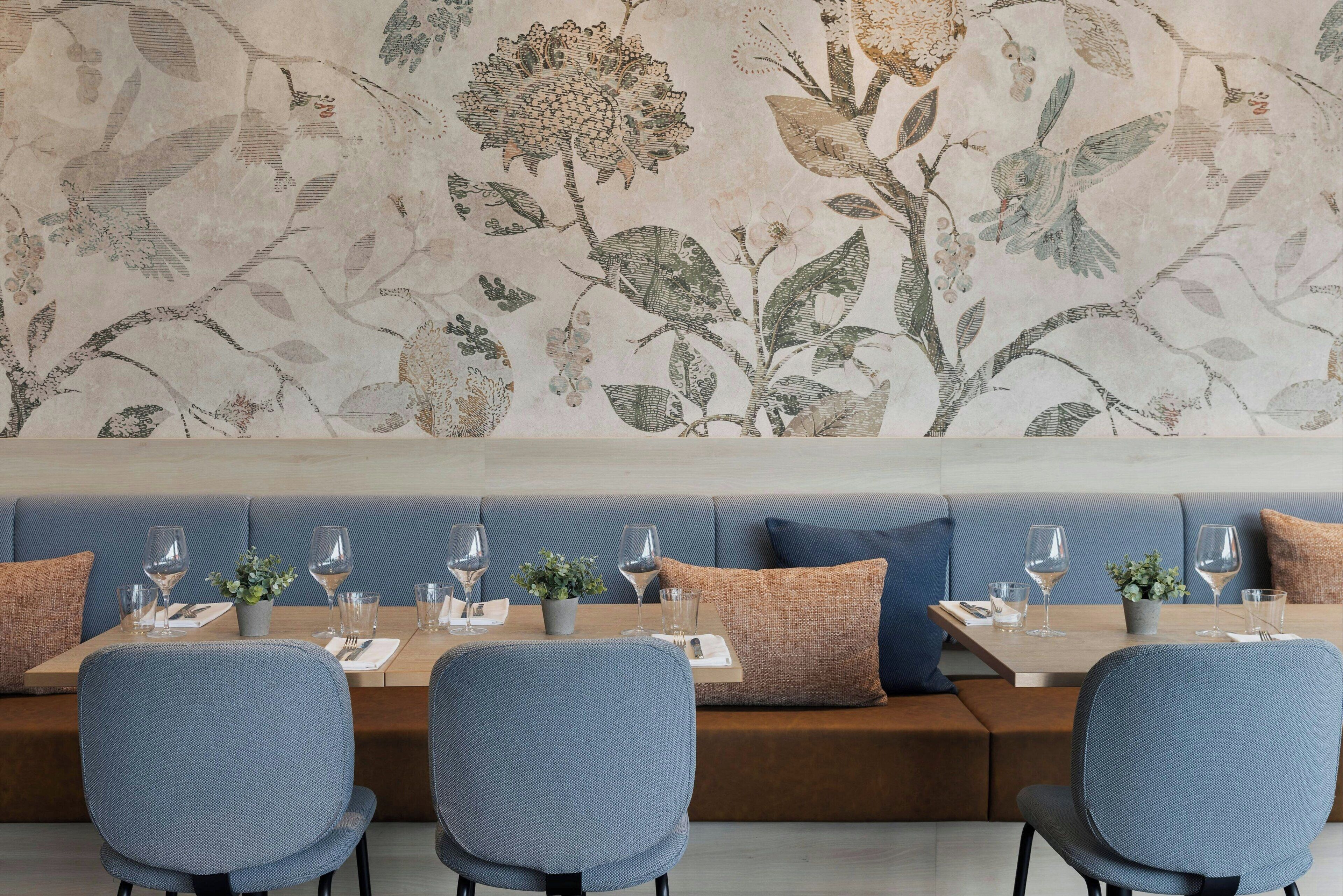 A dining area with a long table set for six, blue upholstered chairs, and a wall with a floral mural.
