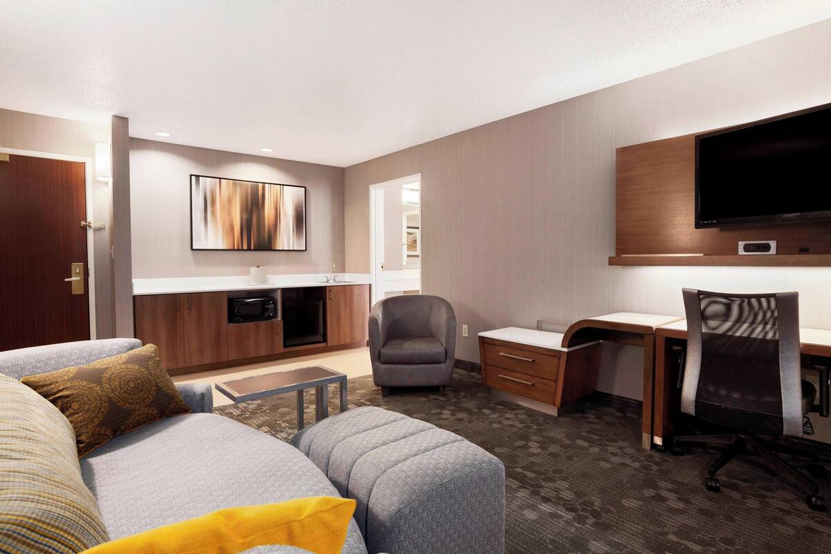 A modern hotel room with a grey sofa, a small table, a desk with a chair, a flat-screen TV mounted on the wall, and a kitchen area with cabinets and a microwave.