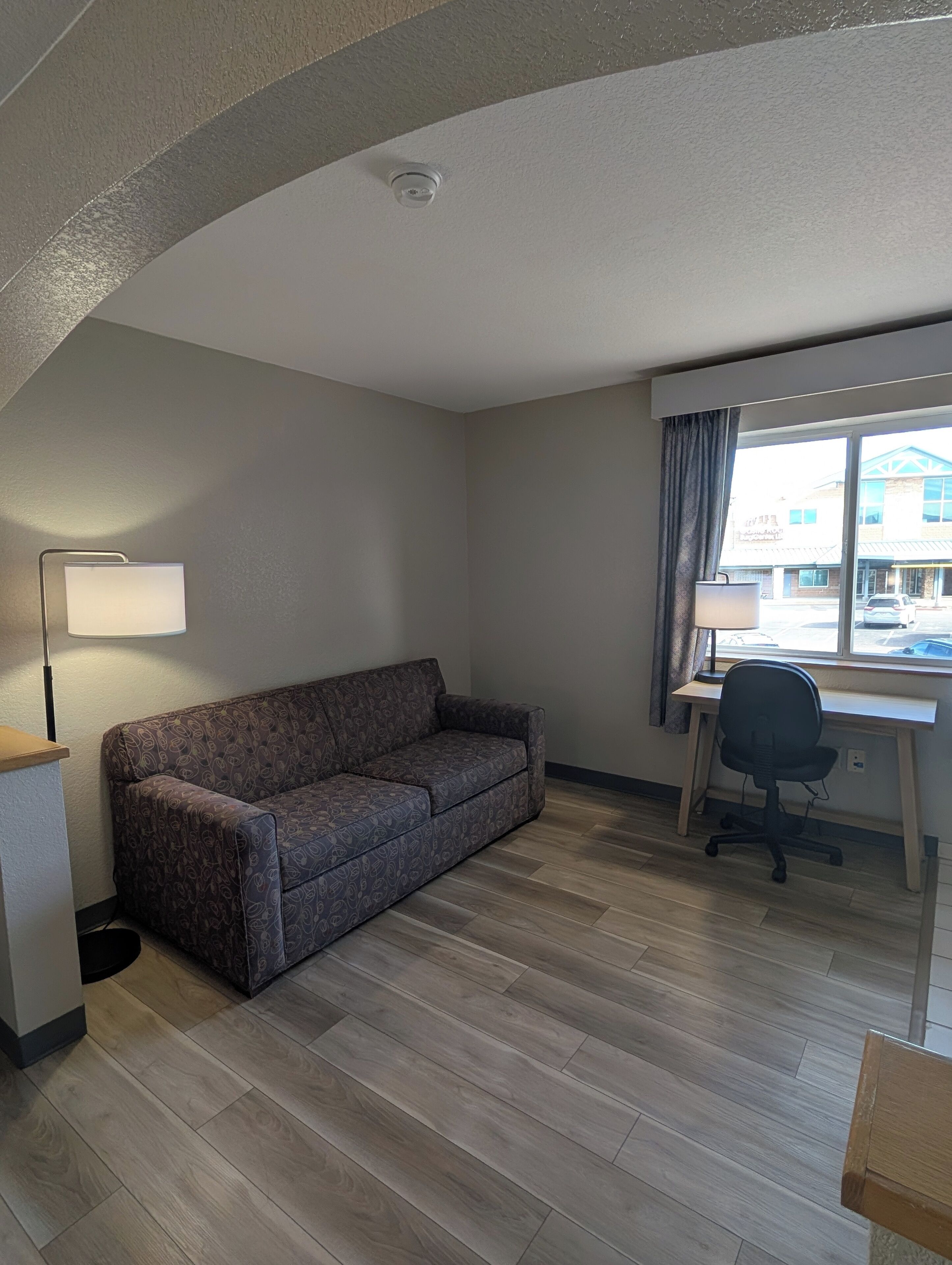 A room with a sofa, a desk with a chair, a lamp, and a window with a view of a building outside.