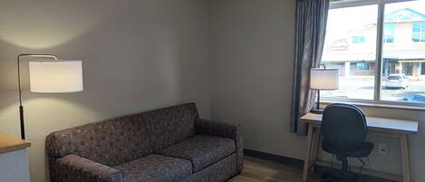 A room with a sofa, a desk with a chair, a lamp, and a window with a view of a building outside.