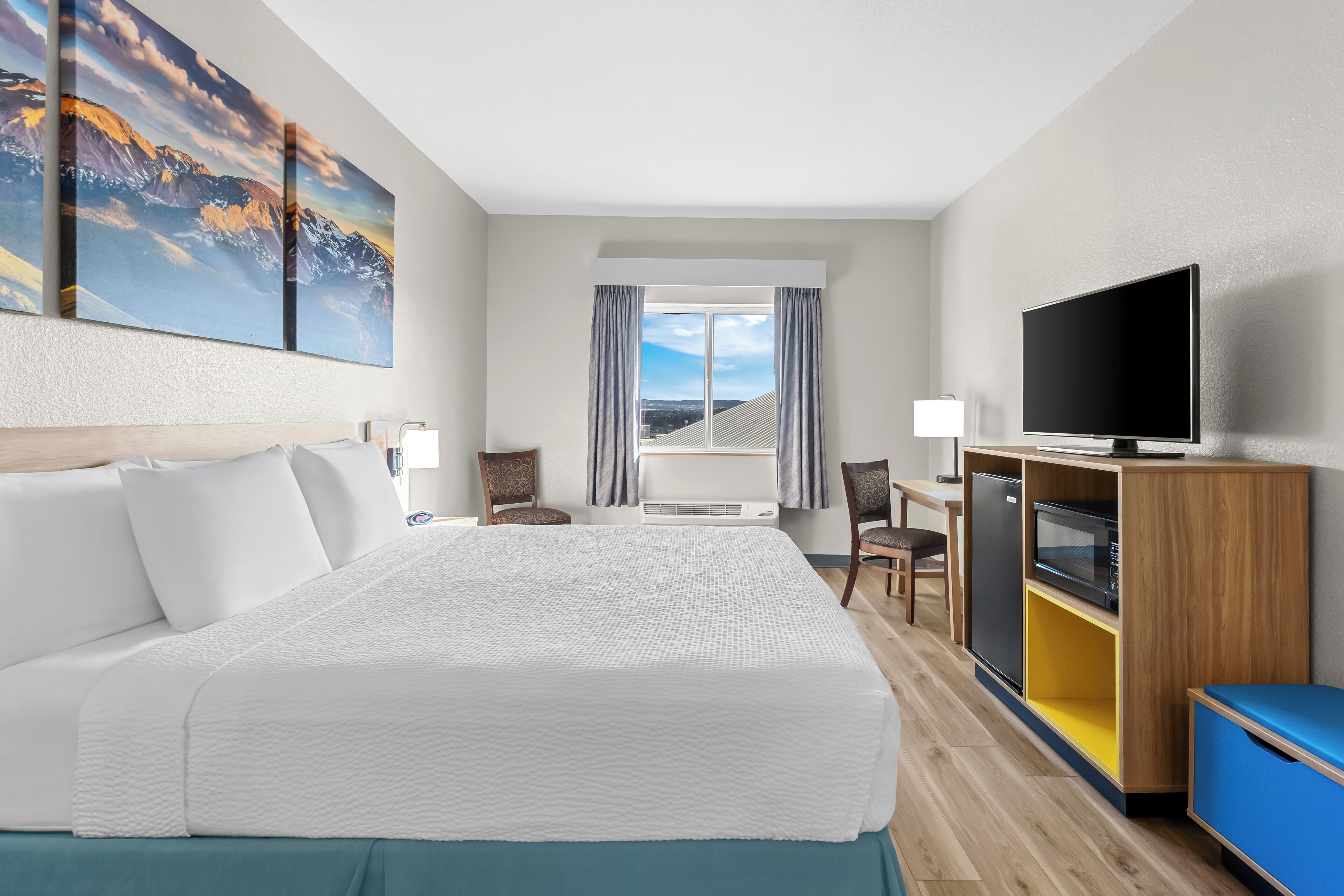 A hotel room with a large bed, a flat-screen TV, a desk with a computer, and a view of the mountains through the window.