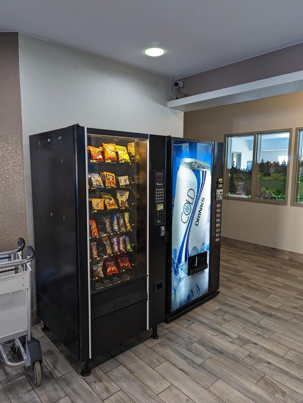A vending machine with snacks and a drink dispenser in a room with wooden flooring and a window with a view of trees. - Days Inn & Suites by Wyndham Castle Rock (Castle Rock)
