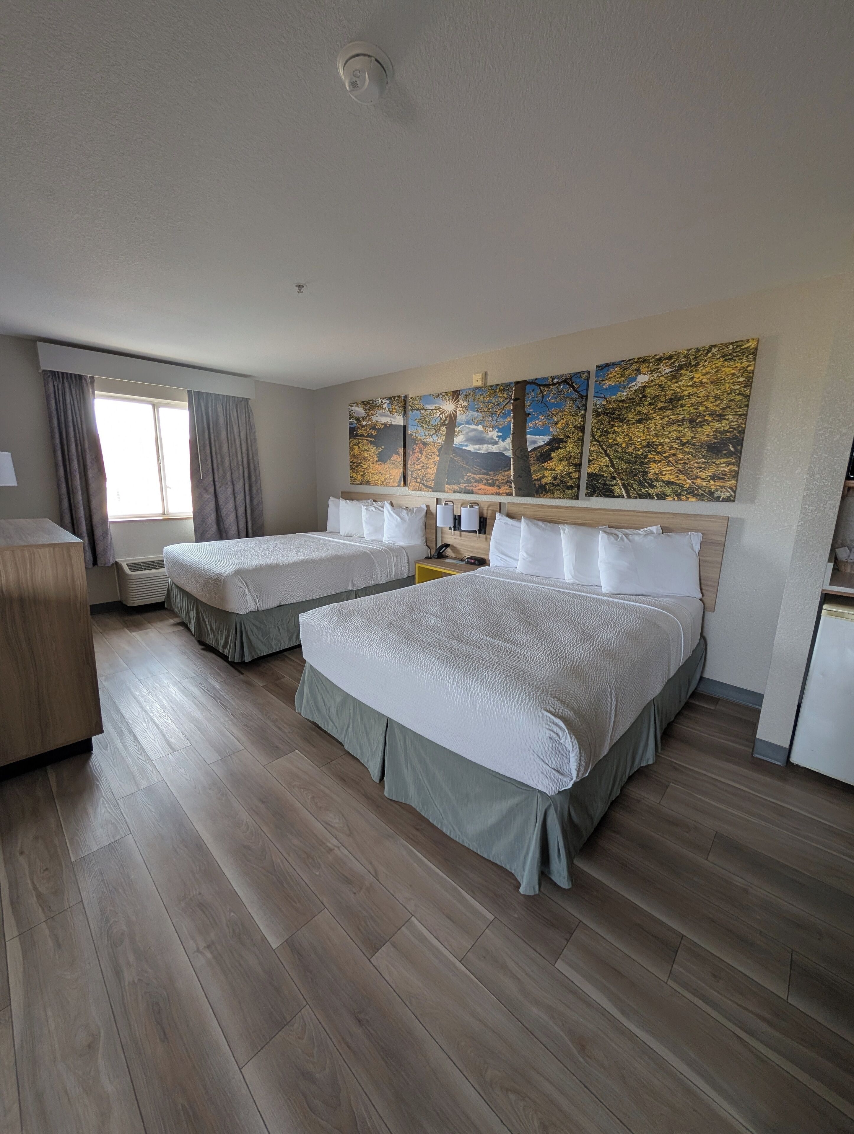 A hotel room with two beds, a desk, a small refrigerator, and a window with curtains.