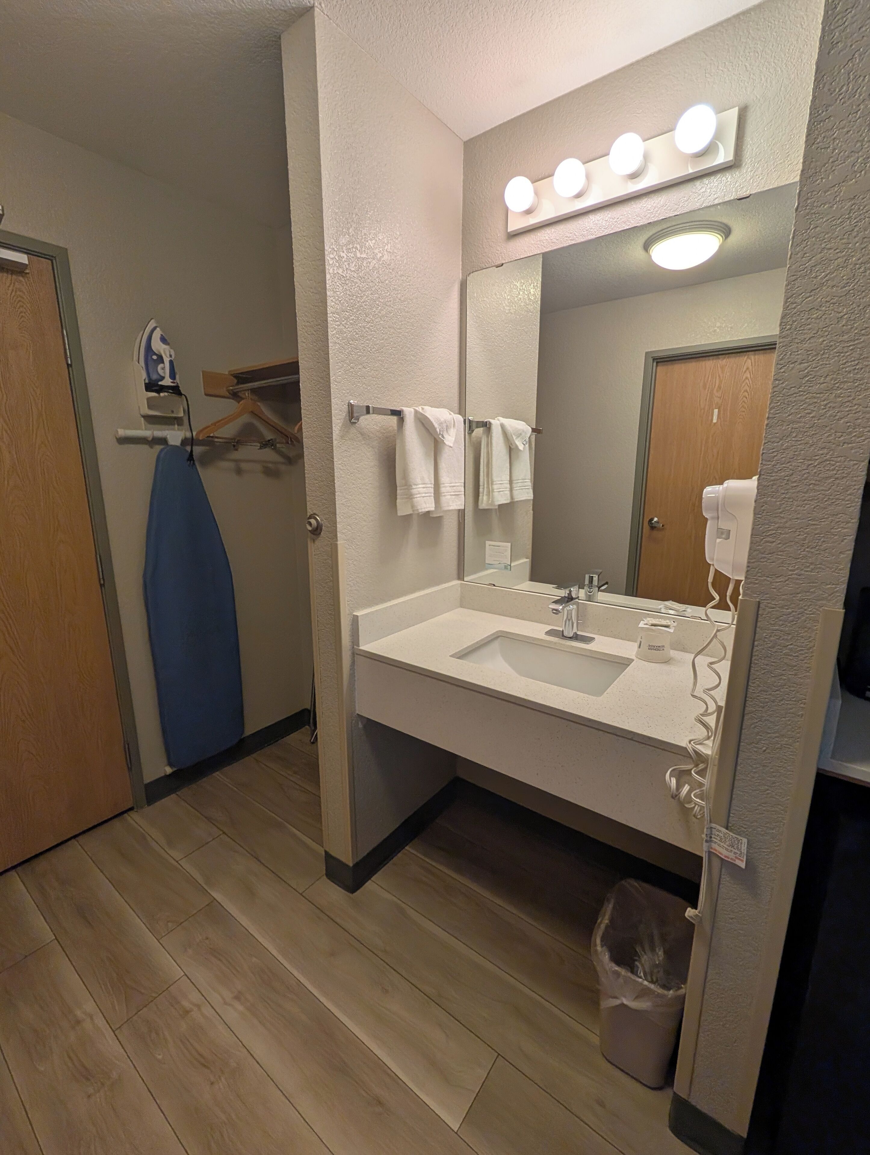 A bathroom with a sink, mirror, and vanity lights. There is a towel rack with towels, a hanging blue garment, and a trash can.