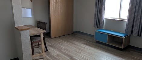 Una habitación compacta con suelo de madera, un escritorio con silla, un armario, un banco y una ventana con cortinas.
