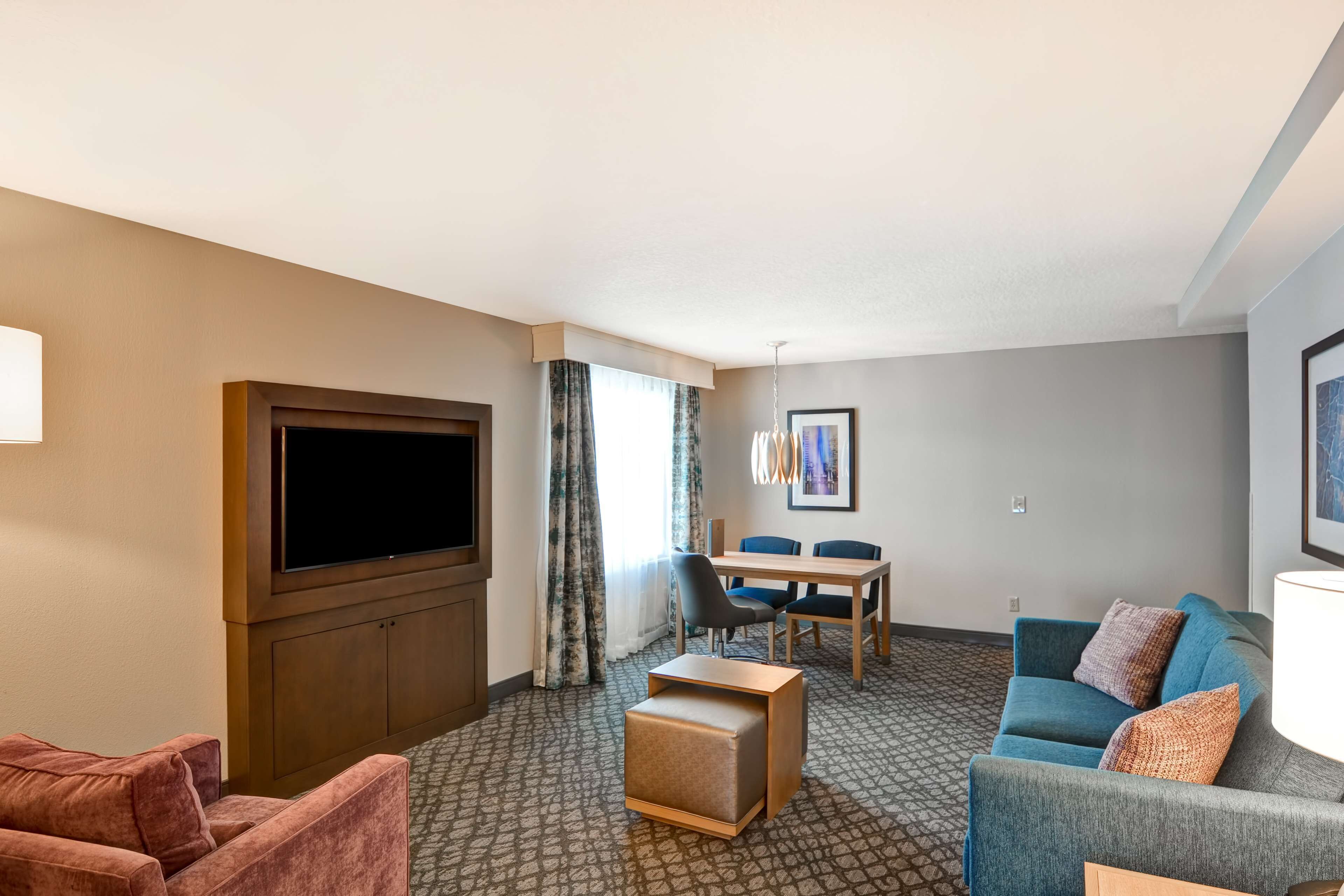 A hotel room with a flat-screen TV, a sofa, a loveseat, a small ottoman, a dining table with chairs, and a patterned carpet.