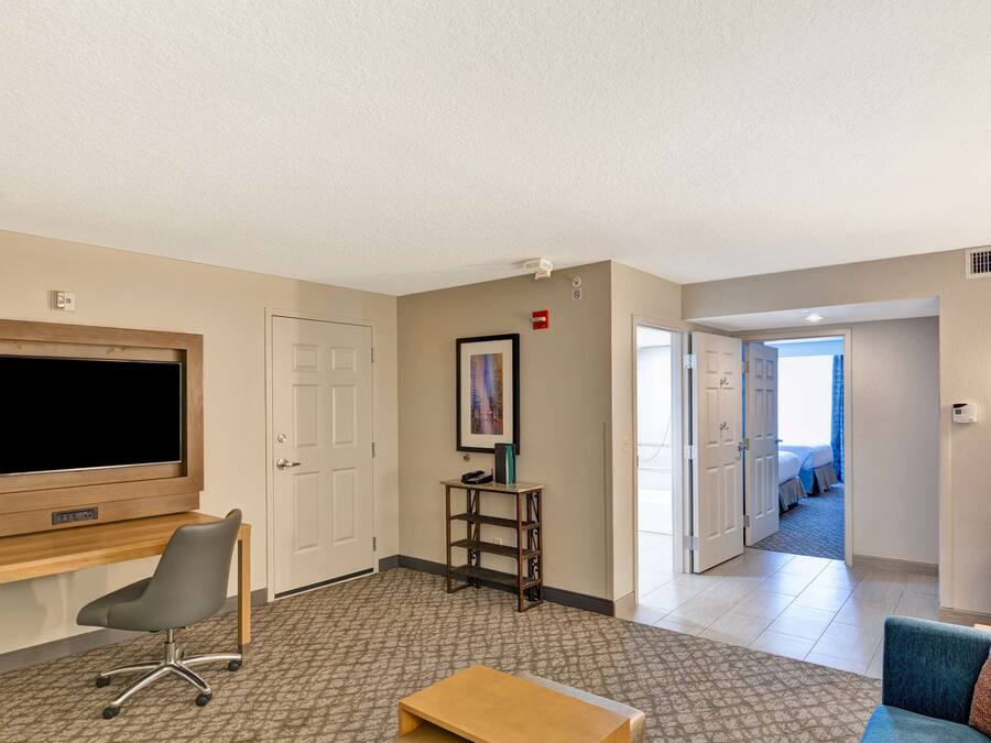 Ein Hotelzimmer mit Flachbildfernseher, Schreibtisch, Stuhl, kleinem Tisch, einem gerahmten Bild an der Wand und einer Tür zum Schlafzimmer.