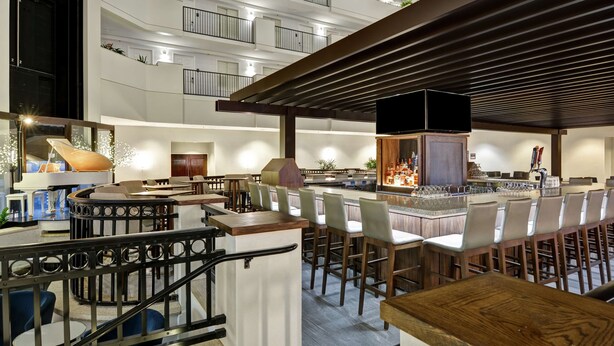 Zona de bar moderna con techos altos, varios niveles y diferentes opciones de asientos, incluyendo taburetes y mesas.