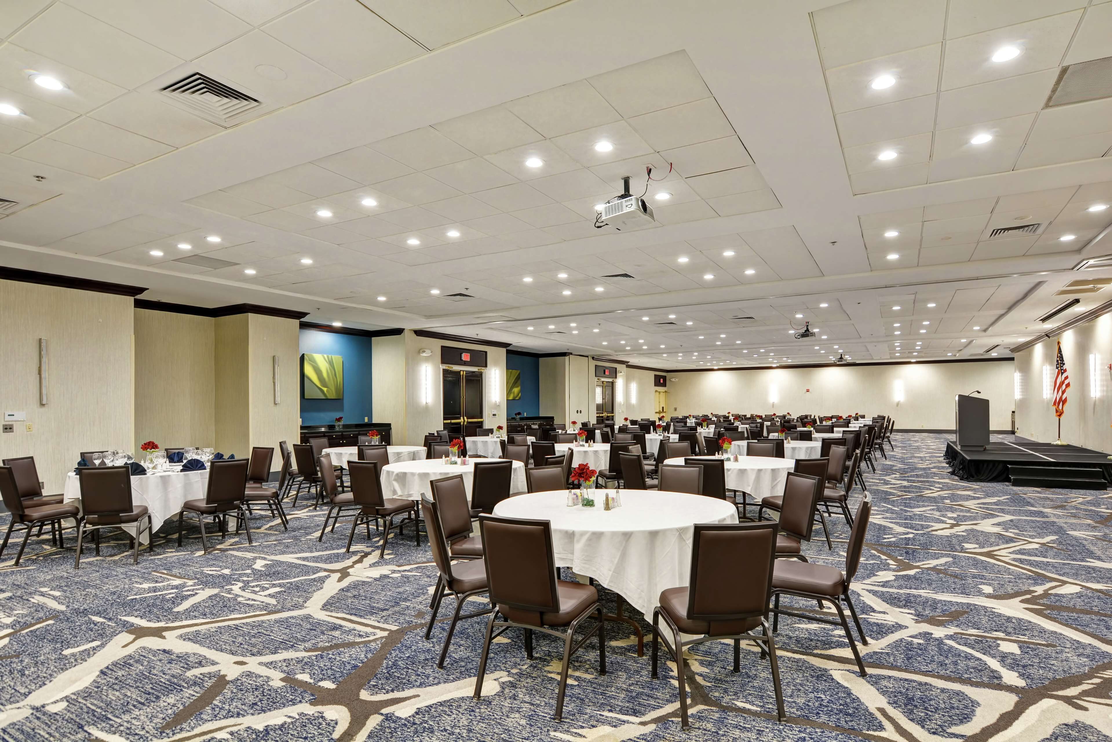 A large conference room with multiple round tables and chairs, a stage, and a carpeted floor.