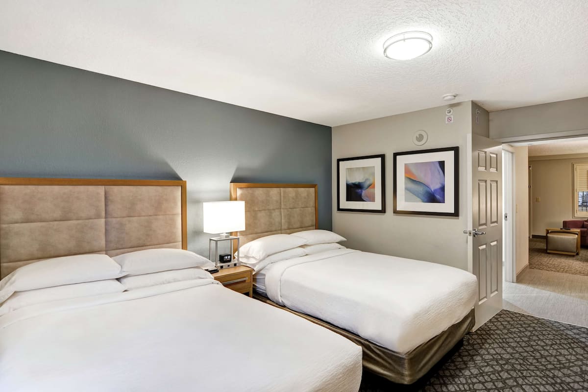 A hotel room with two beds, a bedside table with a lamp, and a door leading to another room.