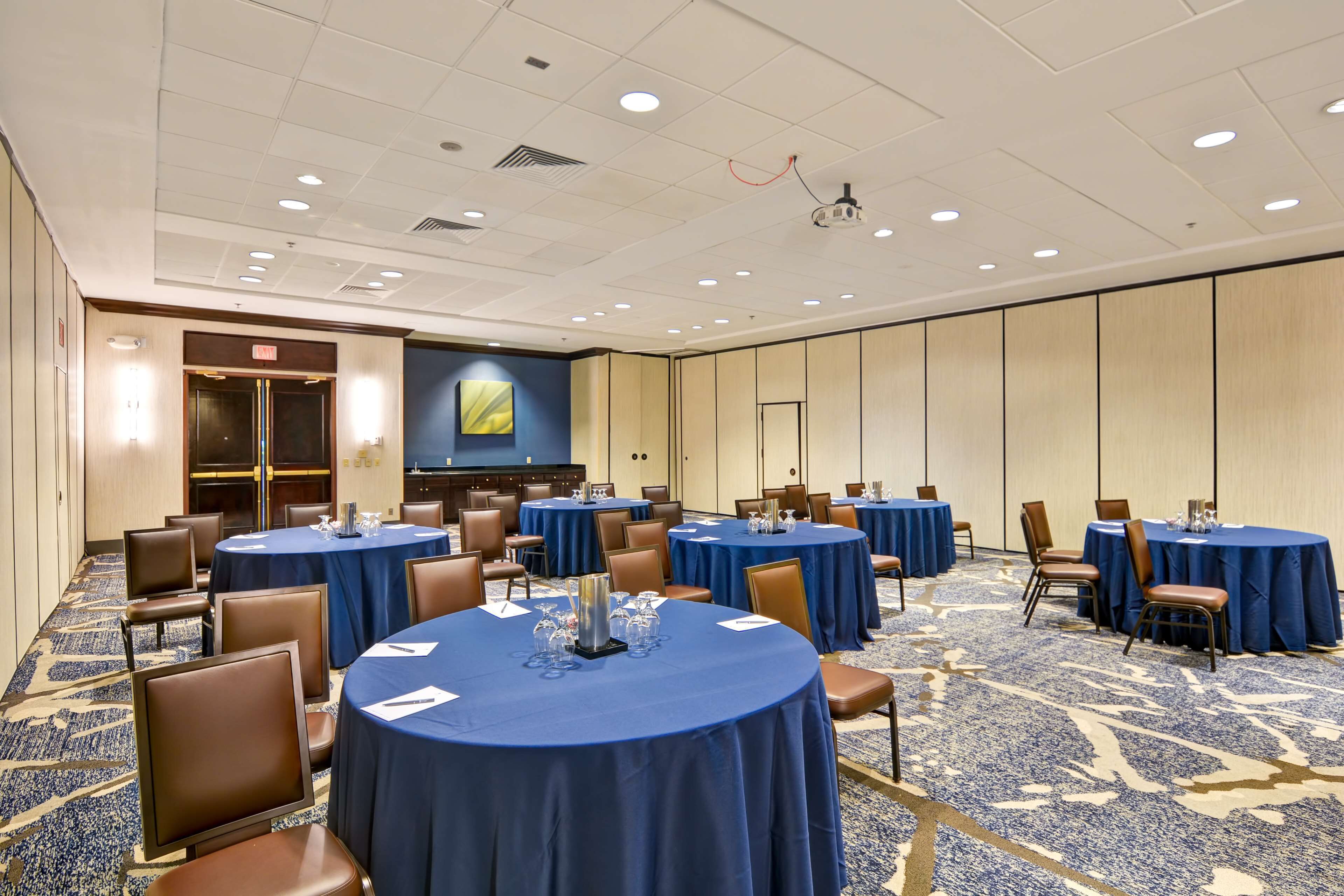 A conference room with multiple round tables covered with blue tablecloths, brown chairs, and a carpeted floor.