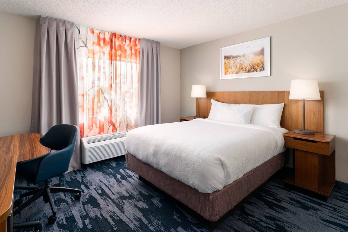 A hotel room with a bed, two bedside lamps, a desk with a chair, a window with curtains, and a wall-mounted picture.
