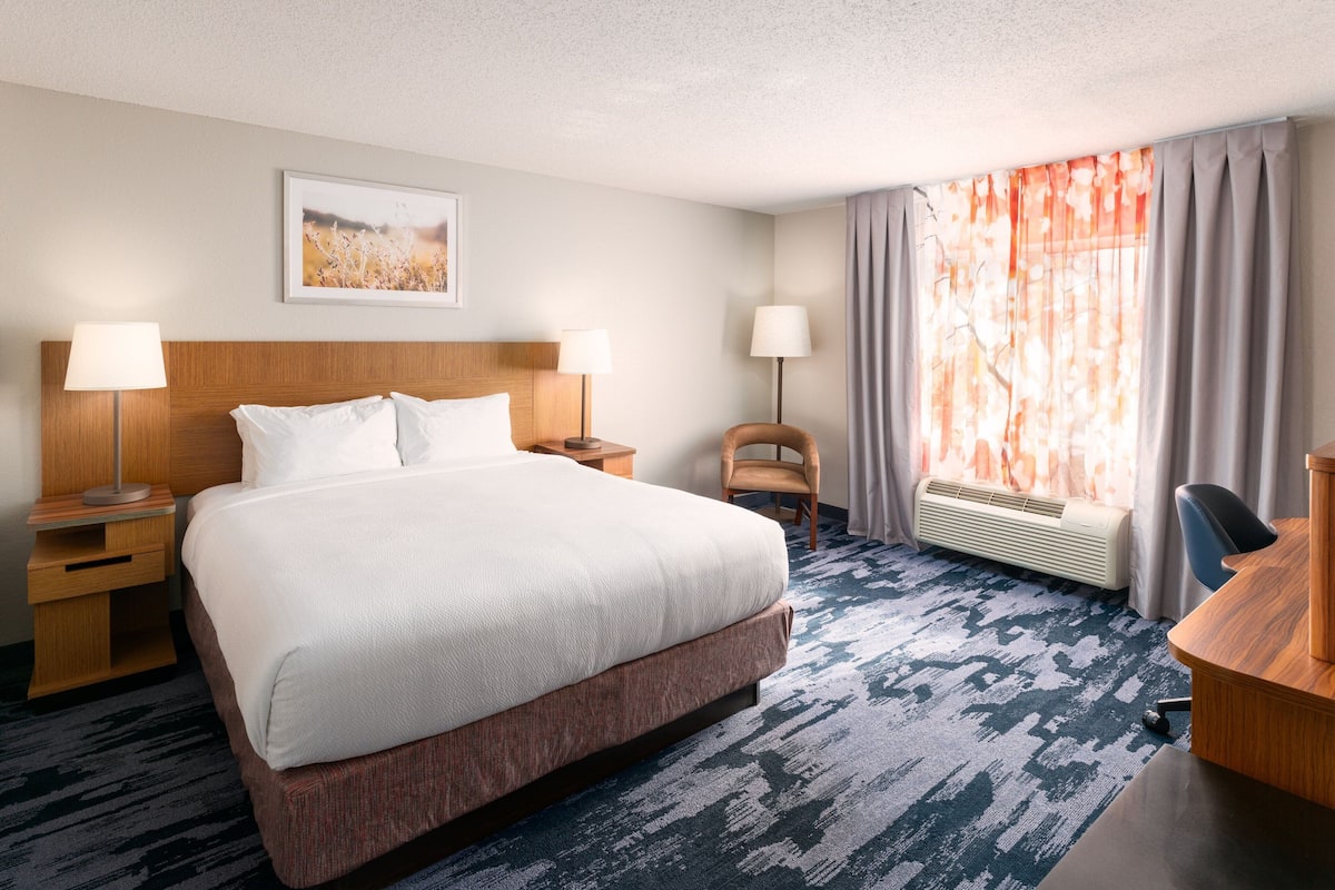 A hotel room with a large bed, two bedside tables with lamps, a desk, a chair, a radiator, and a window with curtains.