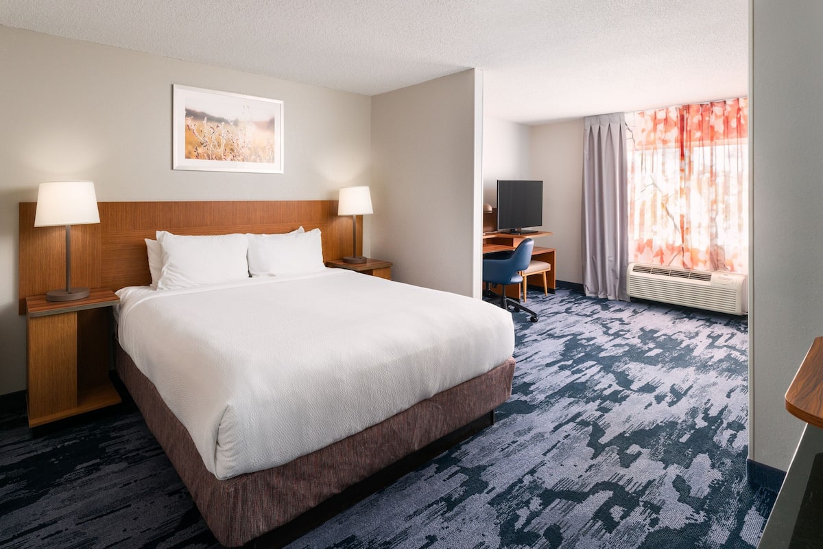 A hotel room with a large bed, two bedside tables with lamps, a desk with a chair, a television, and a window with curtains.