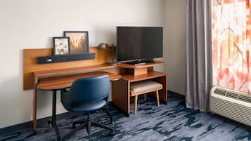 Habitación de hotel con un escritorio de madera, una silla de oficina azul, un televisor de pantalla plana, un pequeño banco y una ventana con cortinas.