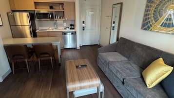 Una sala de estar moderna con un sofá gris, una mesa de centro de madera, y un comedor con sillas, con una cocina al fondo.