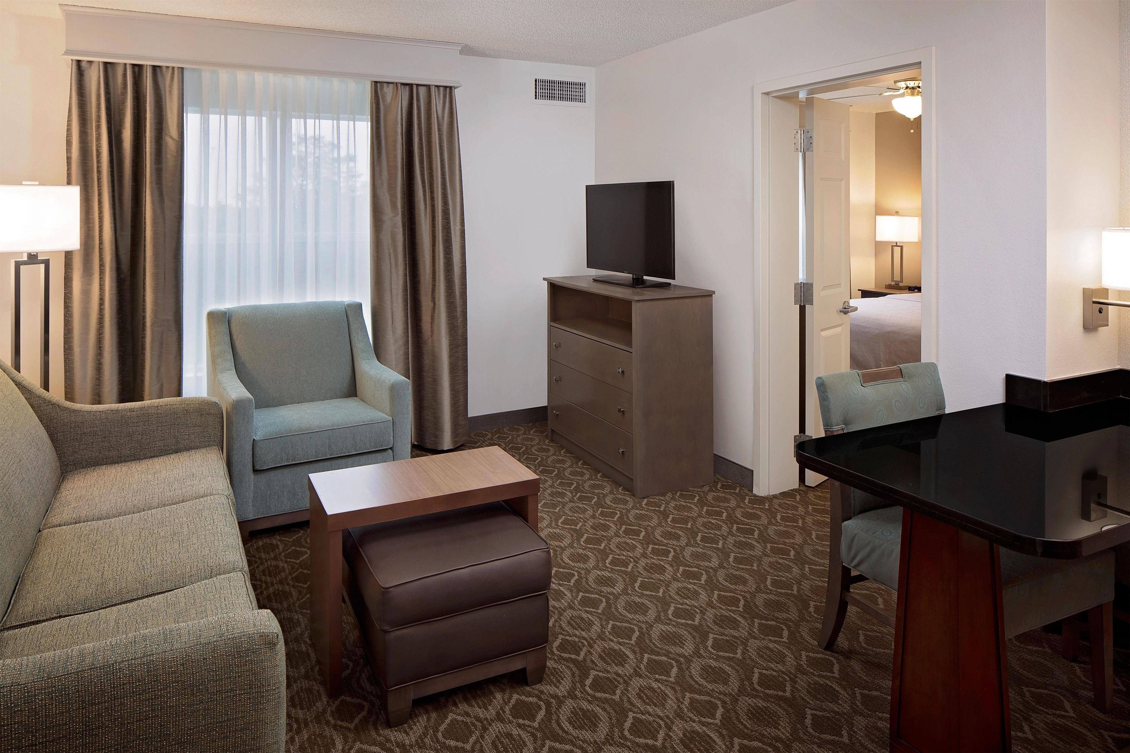 A hotel room with a sofa, a chair, a small table, a TV on a wooden stand, a desk with a chair, a patterned carpet, and a door leading to a bathroom.