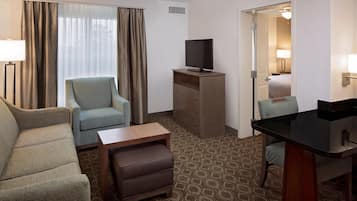 A hotel room with a sofa, a chair, a small table, a TV on a wooden stand, a desk with a chair, a patterned carpet, and a door leading to a bathroom.
