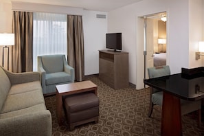 Habitación de hotel con sofá, sillón, una mesita, televisor sobre un mueble de madera, escritorio con silla, alfombra estampada y puerta que da a un baño.