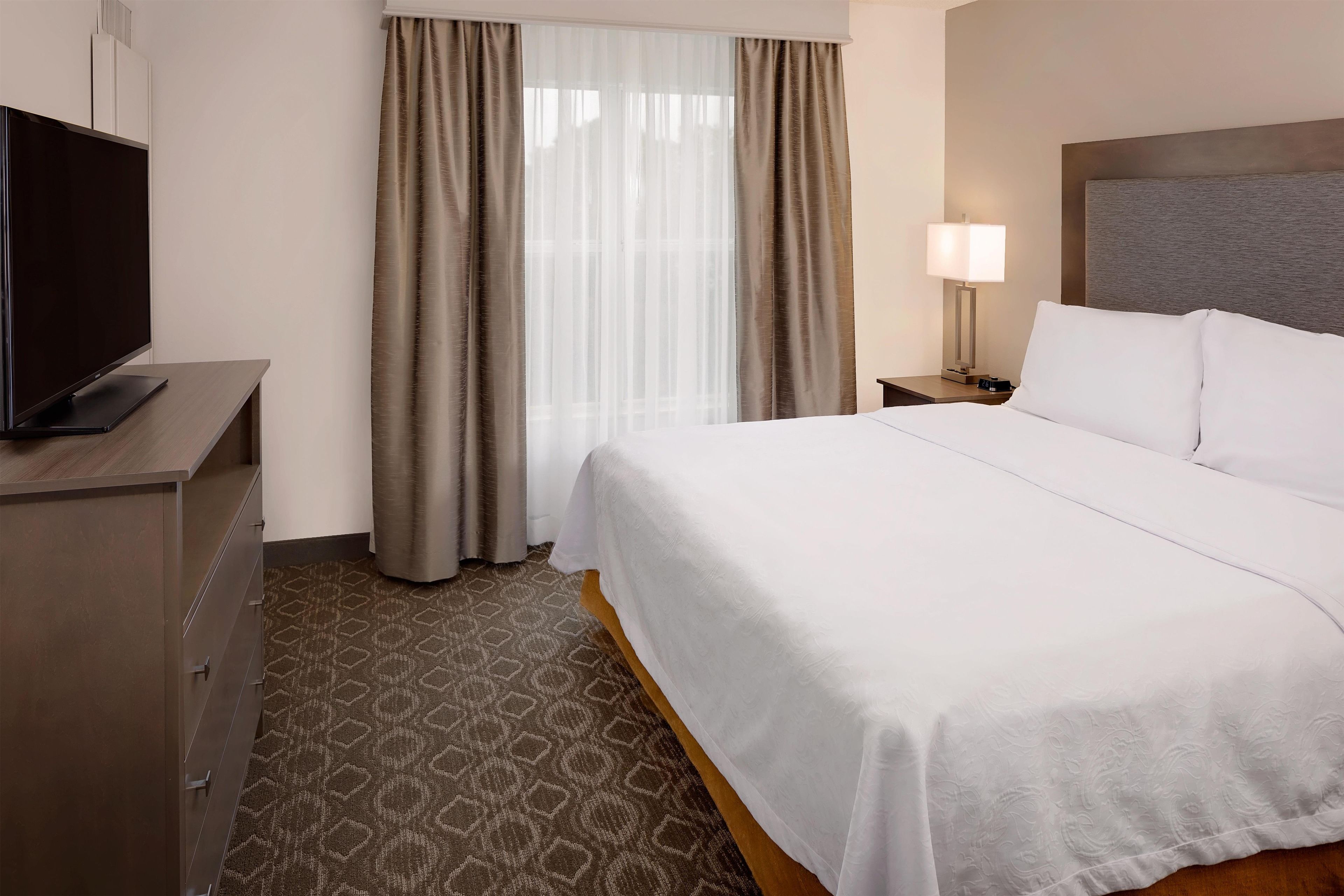 Een hotelkamer met een bed, een tv op een dressoir, een nachtkastje met een lamp, en een raam met gordijnen.