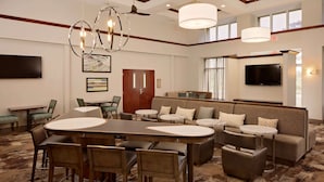 A hotel lobby with a large oval table, several sofas, and chairs. There are ceiling fans, a TV, and framed artwork on the walls. - Homewood Suites by Hilton Orlando-Maitland (Maitland)